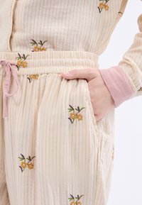 Crème-kleurige, gestreepte stof met bloemenborduursel. Heeft een elastische tailleband, zakken en zachte roze accenten op de mouwen.