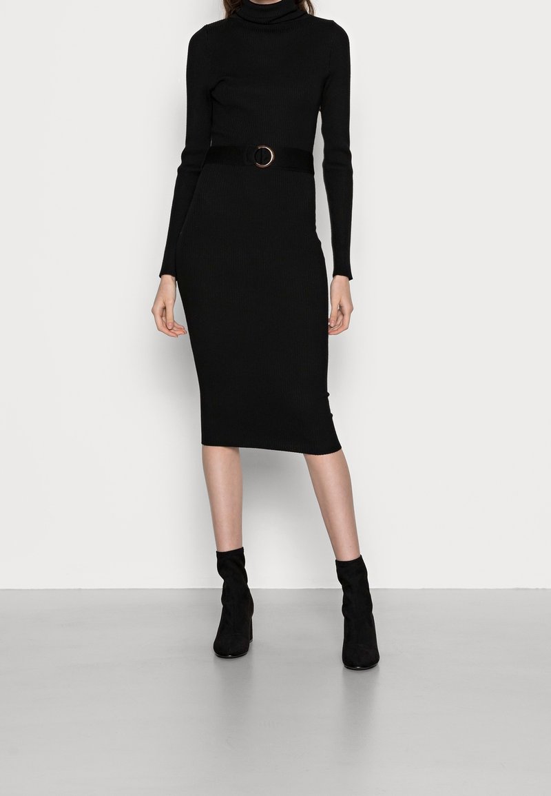 Zwarte geribbelde gebreide jurk met een hoge hals, lange mouwen en een getailleerd silhouet, geaccentueerd met een zwarte riem met een ronde gesp.