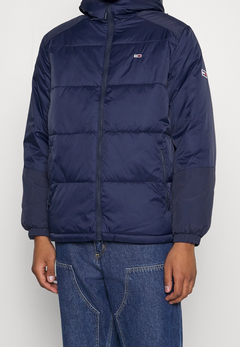 Marineblått vadderat jacka med dragkedja framtill, elastiska ärmslut och logotypdetalj på bröstet och ärmen. Har en huva och en kviltad design.