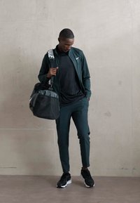 Mand i sort træningstøj og sneakers, der holder en sort duffelbag, står op ad en ensfarvet beige væg og ser nedad.