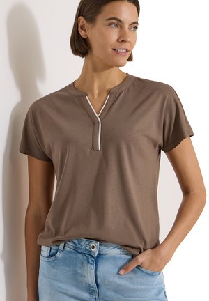 Femme aux cheveux bruns courts portant un haut marron à manches courtes avec un col en forme d’encoche et un jean bleu clair, une main dans la poche.