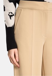 Pantalon beige sur mesure avec plis à l'avant, confectionné dans un tissu lisse. Manche de pull noir avec un motif floral blanc visible sur le côté.