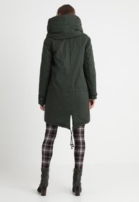 Žena stojící zády, oblečená v tmavě zelené kapucové parce, černobílých kostkovaných legínách a tmavě šedých nároční botách na podpatku.