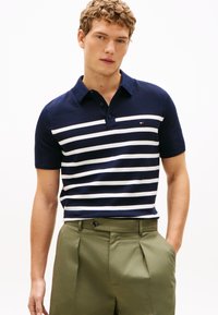 Marineblå og hvide stribede kortærmede polo med krave, der har en lille logo på brystet, parret med lysegrønne skræddersyede bukser.