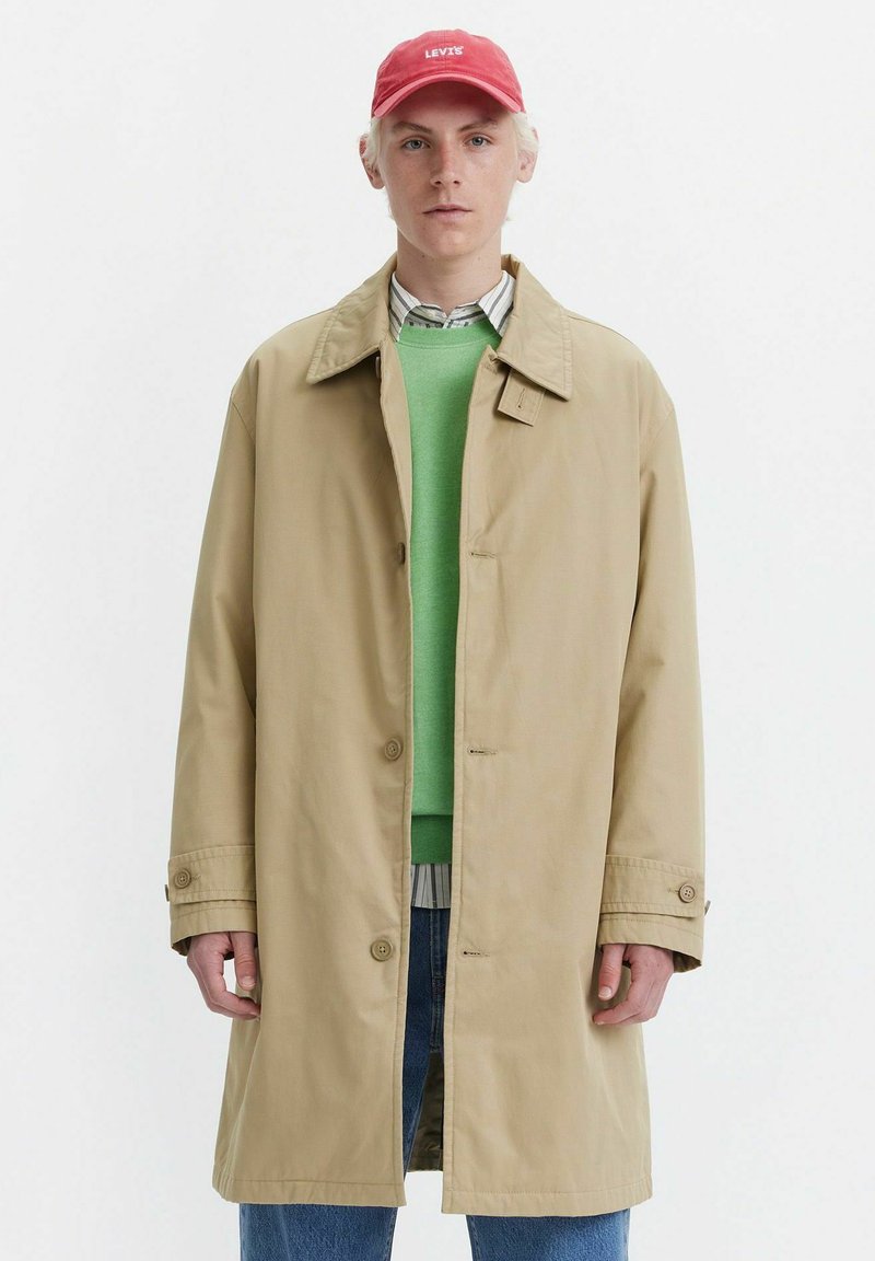 Trench coat beige con una vestibilità comoda, realizzato in tessuto leggero. Indossato sopra un maglione verde, con un cappellino rosso con logo bianco.