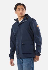 Młody mężczyzna w granatowej kurtce z kapturem, zapinanej na guziki z przednimi kieszeniami oraz w niebieskich dżinsach, patrzący na bok na białym tle.