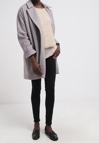 Grå ullkappa med kellerade slag, bärs över en texturerad beige tröja, ihop med svarta skinny jeans och loafers. Enkel och modern design.