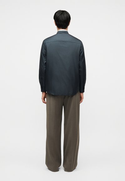 Άνδρας που στέκεται με την πλάτη γυρισμένη, φοράει σκούρο μπλουζάκι μακρυμάνικο και καφέ χαλαρό παντελόνι, μπροστά σε λευκό φόντο.