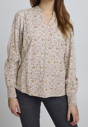 Kobieta ubrana w bluzkę z długim rękawem w różowy, zielony i beżowy wzór kwiatowy oraz czarne dżinsy, stojąca na tle gładkiego szarego tła.