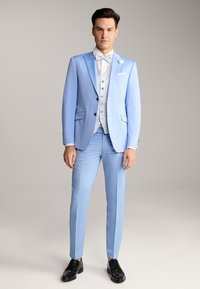 Hellblauer Anzug mit passender Jacke und schmal geschnittenen Hosen. Dazu eine weiße Weste, eine Fliege und schwarze Schuhe. Subtile Texturdetails.