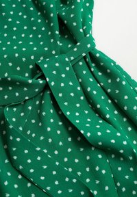 Tissu vert à pois blancs, avec un détail de taille nouée. Le matériau semble doux, avec une texture lisse et un design fluide.