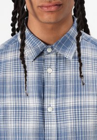 Blauw-wit geruite button-up shirt met een klassieke kraag, voorzien van een zachte textuur en plastic knopen. Het ontwerp bevat horizontale en verticale lijnen.