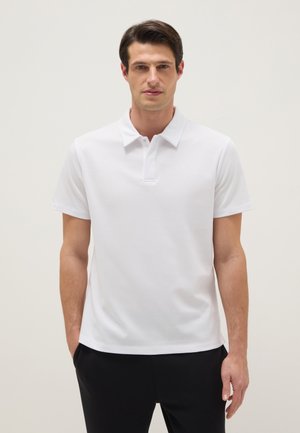Homme debout les mains dans les poches, portant un polo blanc uni à manches courtes et un pantalon noir, sur un fond neutre.