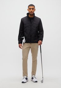 Άνδρας που φοράει μαύρο μπουφάν και μπεζ παντελόνι κρατάει μπαστούνι γκολφ και λευκό γάντι σε λευκό φόντο.