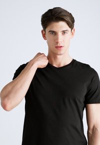 Zwarte V-hals t-shirt van een zachte, gladde stof. Aansluitend ontwerp met korte mouwen. Het model poseert met één hand op zijn schouder tegen een lichte achtergrond.