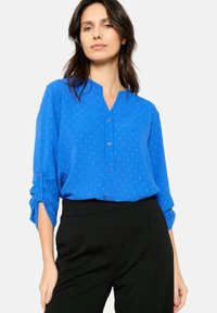 Camicia blu con colletto alla mandarino, caratterizzata da un sottile motivo texturizzato e maniche arrotolabili, abbinata a pantaloni neri a vita alta.