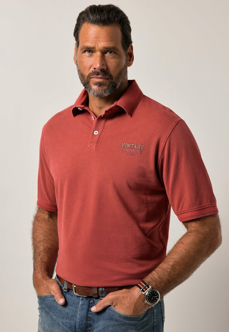 Polo shirt rosso con colletto e maniche corte. Presenta il logo "VINTAGE DENIM" in un colore a contrasto. Indossato con jeans blu e un orologio.