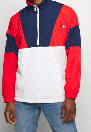 Persona con una chaqueta Adidas roja, azul marino y blanca con cremallera frontal y vaqueros azul claro, de pie frente a un fondo liso.