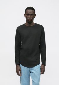 Mladý muž s tmavou pletí, oblečený v jednoduchém černém tričku s dlouhým rukávem a světle modrých džínech, stojící před jednobarevným bílým pozadím.