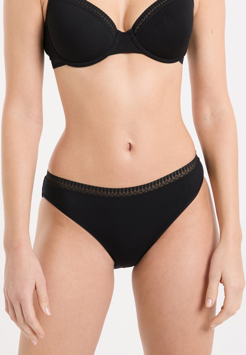 Donna che indossa un reggiseno nero e slip neri coordinati con rifiniture decorative, in piedi contro uno sfondo bianco.