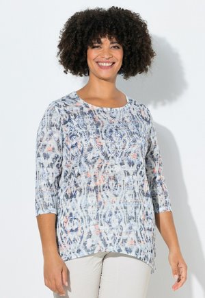 Smilende kvinde med krøllet hår iført en lys mønstret langærmet top og lyse bukser, stående mod en hvid baggrund.