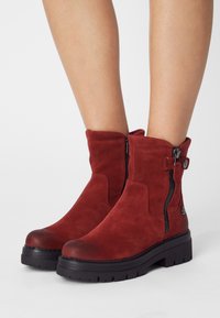 Botines de ante rojo hasta el tobillo con punta redonda, cremalleras laterales y una suela texturizada negra. Presenta un detalle decorativo en forma de botón en el lateral.