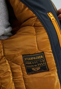 Interno di giacca navy con fodera trapuntata dorata, caratterizzato da un'etichetta nera con la scritta "STEAPACKER ISSUE 409/32" e dettagli in zip gialli.