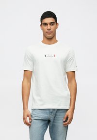 Λευκό βαμβακερό μπλουζάκι με κοντά μανίκια, με ένα ορθογώνιο λογότυπο σε κόκκινο, μαύρο και μπλε στο κέντρο. Συνδυάζεται με ανοιχτό μπλε τζιν.