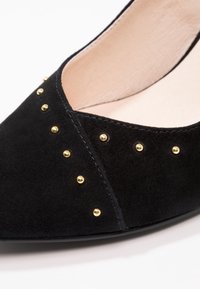 Chaussure plate noire en suède à bout pointu avec de petites clous ronds dorés disposés en diagonale le long du bord supérieur et du cou-de-pied.