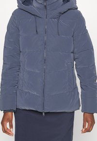 Navy blue puffer jacket with a zip front, hood, and adjustable drawstrings. Features horizontal stitching and a front pocket.