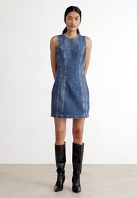 Jeanskjole uten ermer med en tettsittende silhuett, prydet med dekorative nagler, blå farge og nagleakkenter langs sømmene.