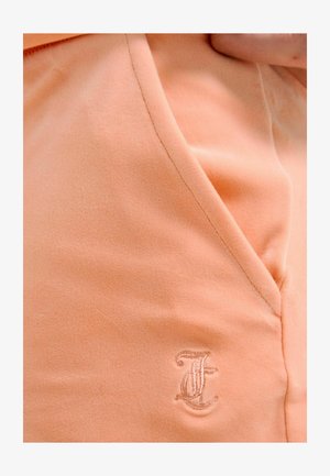 Close-up van perzikkleurige stof met een gestikte naad en geborduurde letter "J" nabij de rand van een kledingstuk.