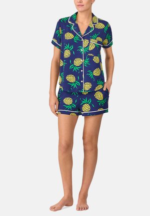 Persona che indossa un pigiama coordinato a maniche corte e pantaloncini con stampa di ananas giallo brillante su tessuto blu scuro, in piedi a piedi nudi.