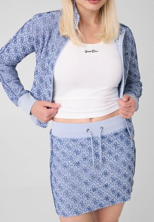 Femme aux cheveux blonds portant un ensemble veste et jupe bleu à motifs avec un crop top blanc, mains tenant les bords de la veste, faisant face à l'avant.