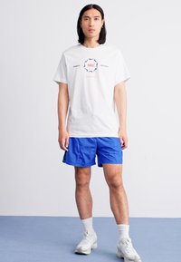 Mand står iført en hvid Nike t-shirt, blå træningsshorts, hvide sokker og hvide sneakers foran en ensfarvet baggrund.