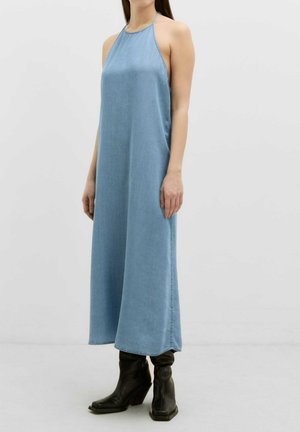 Robe halter en denim bleu clair avec une coupe ample et un design long jusqu'au sol. Présente un col montant et un style minimaliste. Portée avec des bottes à talons noires.