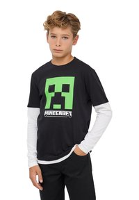 Chłopiec z jasnobrązowymi włosami w czarnej koszulce Minecraft na białych długich rękawach, stojący z jedną ręką w kieszeni na jednolitym tle.