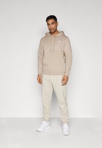 Beige hoodie met een diagonaal textuurpatroon, voorzien van een voorzak en drawstrings, gecombineerd met lichte grijze sweatpants en witte sneakers.