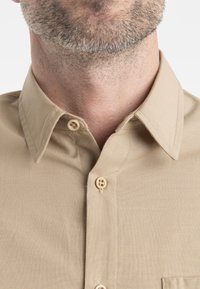 Camicia beige con colletto a punta, dotata di bottoni color nocciola, tessuto texturizzato e una sola tasca sul petto. Design pulito e semplice.