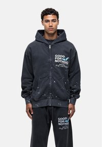 Donkergrijze zip-up hoodie met spetterpatroon en voorzakken. Bevat tekst "GOOD FOR NOTHING" en een vlinderafbeelding. Zachter, katoenen materiaal.