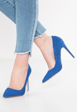 Fødder iført knaldblå ruskind høje hæle kombineret med lyseblå skinny jeans med frynset kant på en hvid gulv.