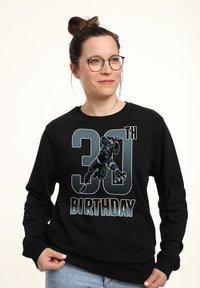 Sudadera negra con un gráfico de un personaje, texto "30 CUMPLEAÑOS" en negrita, con acentos en azul y gris, cuello redondo y mangas largas.