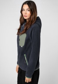 Marine fleece jack met een groene ritszak en accentdetails op de manchetten. Beschikt over een capuchon en een textielstructuur voor warmte. Inclusief zijzak met rits.