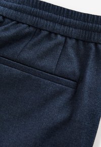Pantalon gris foncé avec une ceinture élastique, une poche passepoilée arrière et un tissu doux et texturé visible en gros plan.