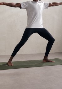 Leggings neri e una maglietta sportiva grigia chiara; la persona è in piedi su un tappetino da yoga verde in posizione guerriero con i piedi nudi.