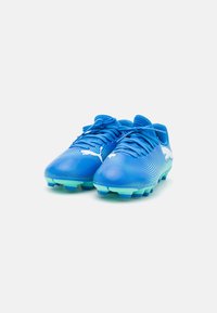 Scarpini da calcio blu con tomaia in mesh texturizzato, caratterizzati da un logo bianco, lacci e un accento verde sulla suola con tacchetti stampati.