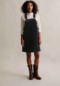 Frau mit lockigem Haar, die ein kariertes Langarmhemd unter einem schwarzen Latzkleid und braunen Lederstiefeln bis zur Mitte der Wade trägt, steht vor einem schlichten Hintergrund.