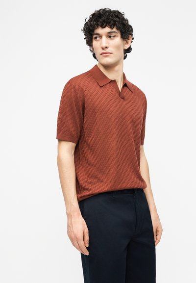 Νεαρός με κατσαρά μαλλιά που φοράει πόλο με υφή σε χρώμα σκουριάς και σκούρο μπλε παντελόνι, κοιτάζοντας προς τα αριστερά του.