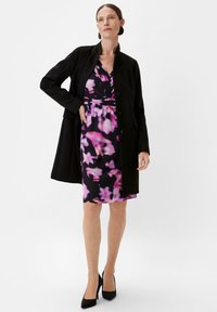Schwarzer Mantel über einem lila floral bedruckten Kleid. Das Kleid hat eine taillierte Taille und einen V-Ausschnitt, kombiniert mit schwarzen hochhackigen Schuhen.
