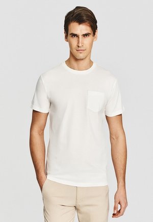 Młody mężczyzna o brązowych włosach, ubrany w gładką białą koszulkę z krótkim rękawem z kieszenią na piersi oraz beżowe spodnie, stoi na jednolitym tle.